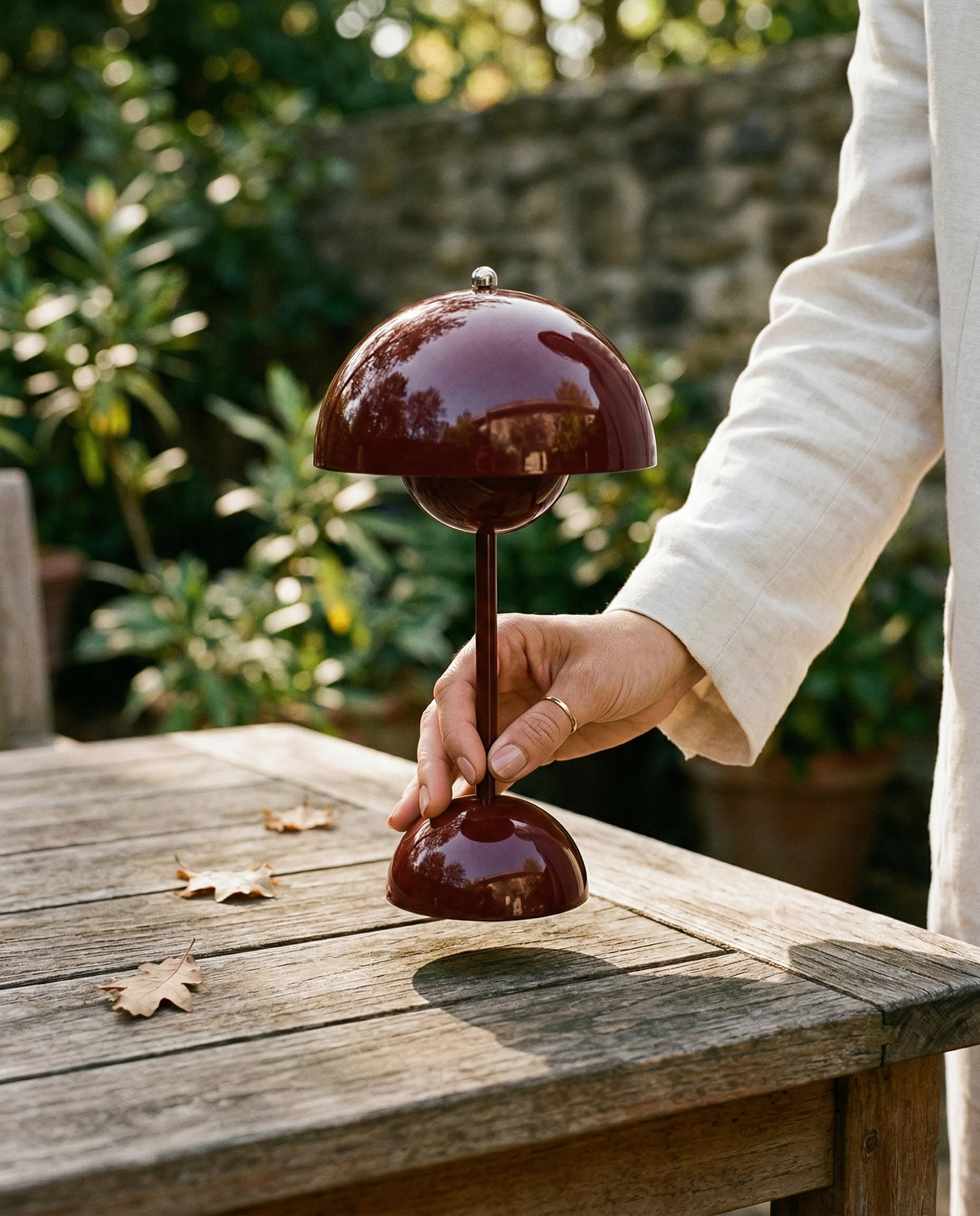 Cordless table lamp DOMA No.02 in wine red held by its stem on a weathered wooden garden table