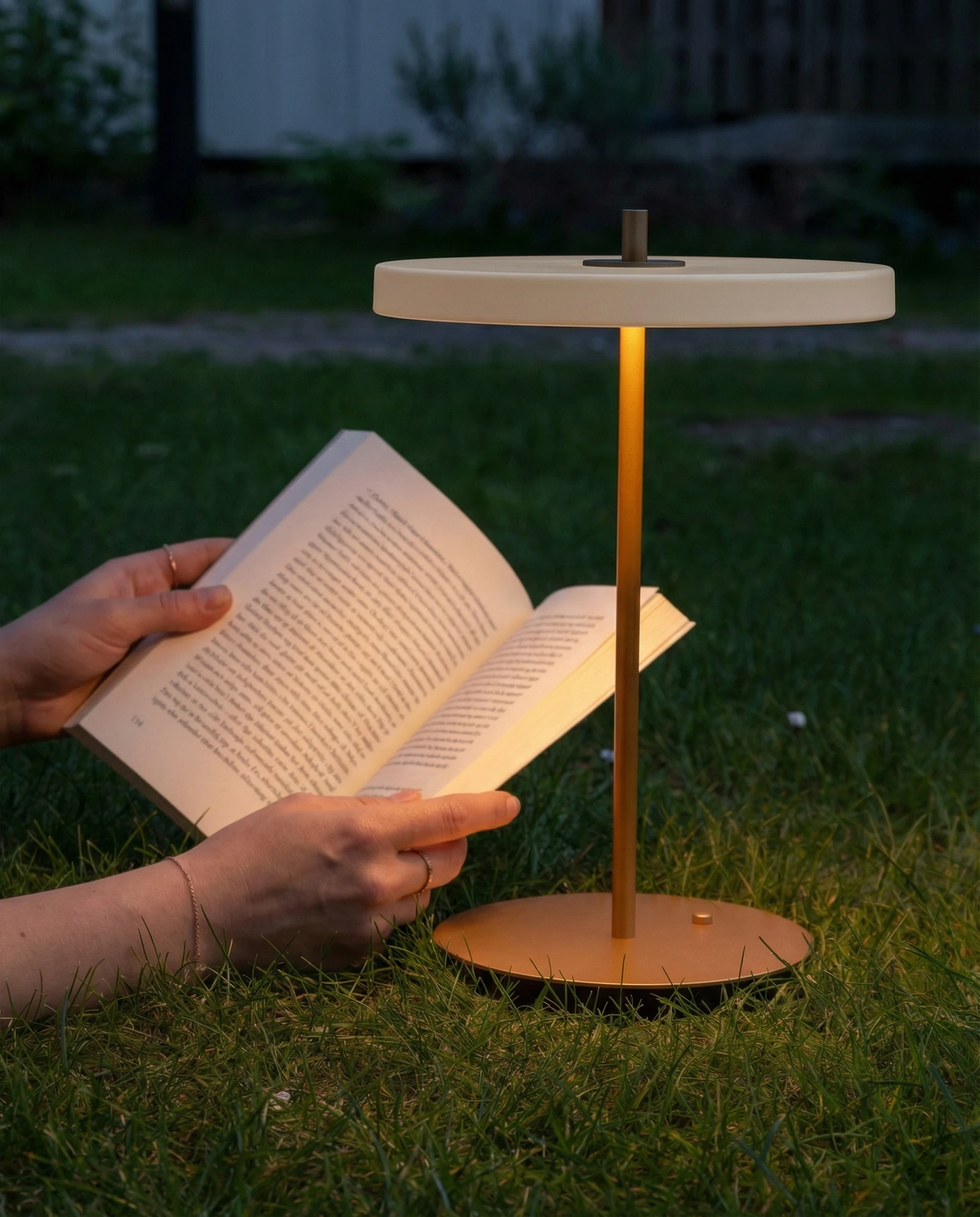 Cordless table lamp SALA No.01 in matte white, lit on a lawn while hands hold an open book beneath it