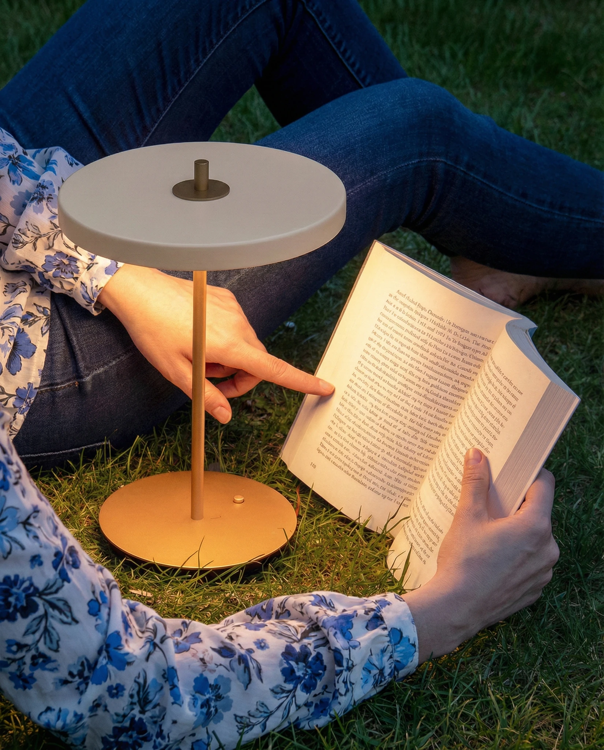 Cordless table lamp SALA No.01 in matte white, lit on the grass beside a woman lying down reading a book