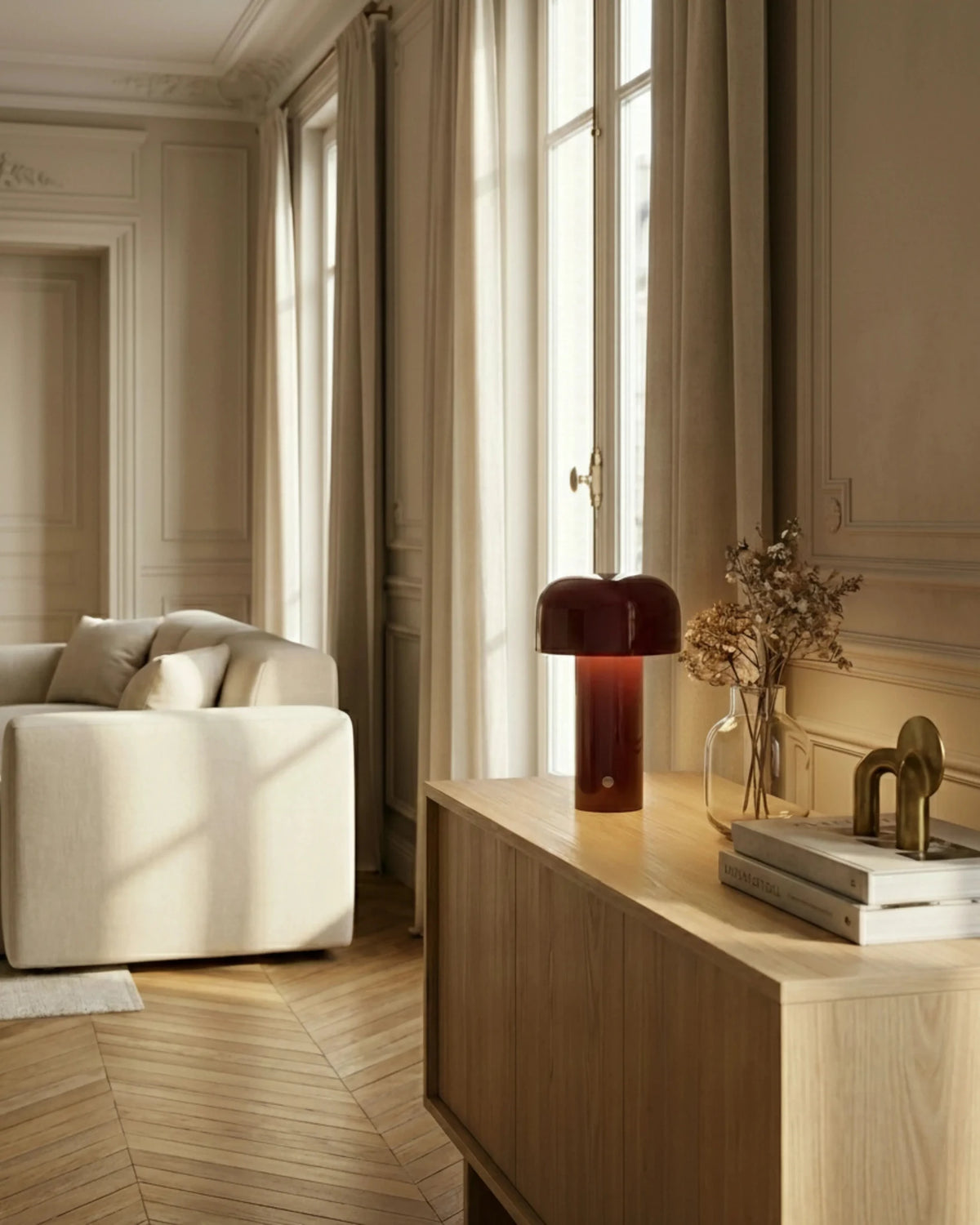 Cordless table lamp DOLCE No.01 in bordeaux red lit on a wooden console with books near a cream sofa