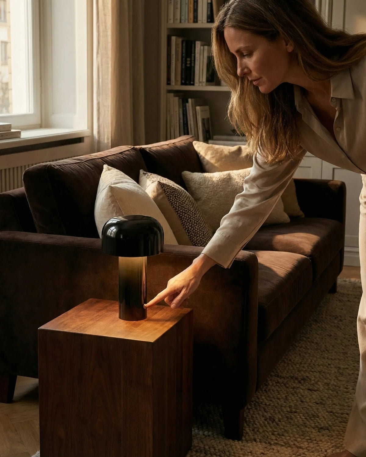 Cordless table lamp DOLCE No.01 in deep black lit on a wooden side table as a woman reaches toward it by a sofa