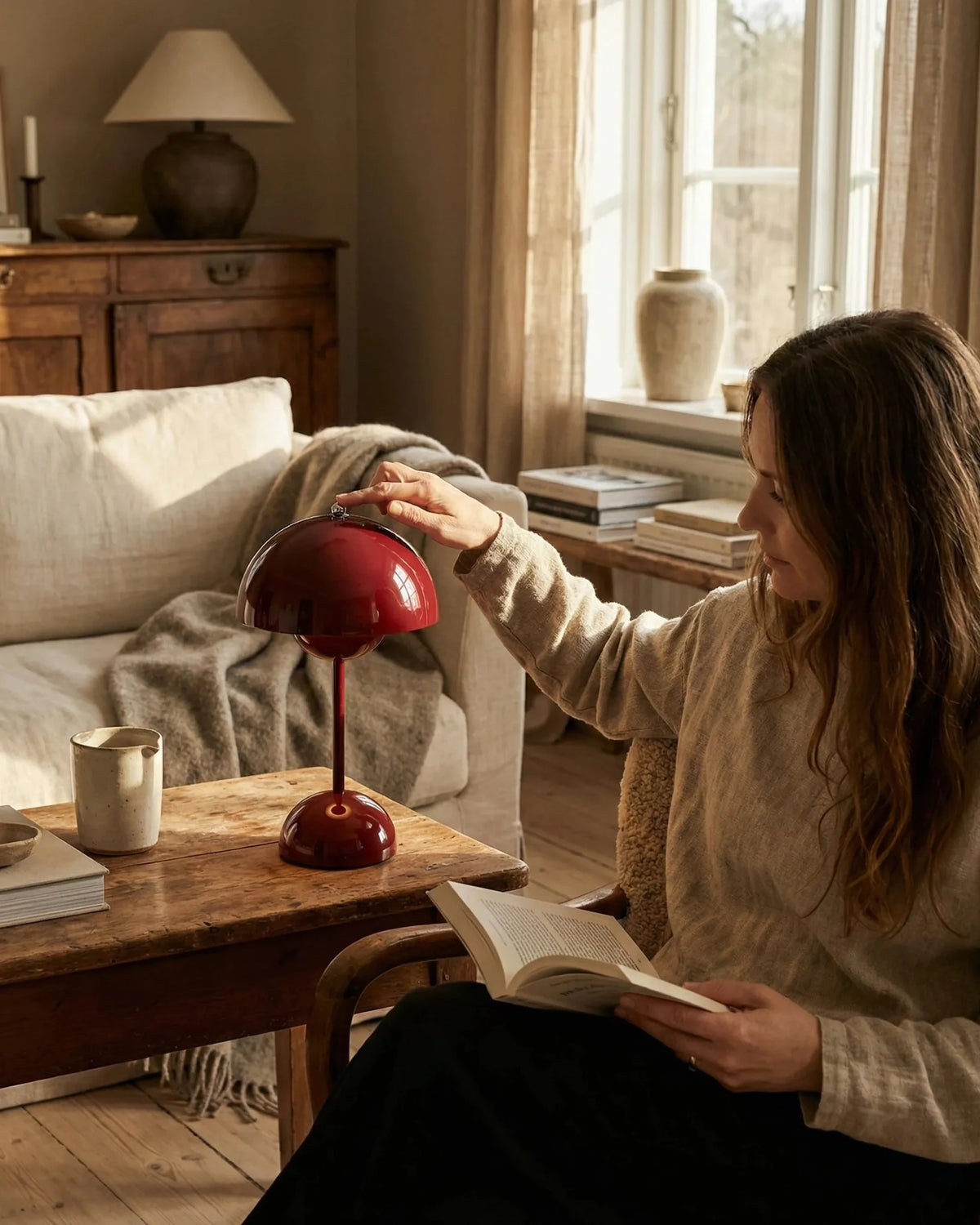 Cordless table lamp DOMA No.02 in wine red on a wooden coffee table, a woman reading on the sofa reaching for the dome