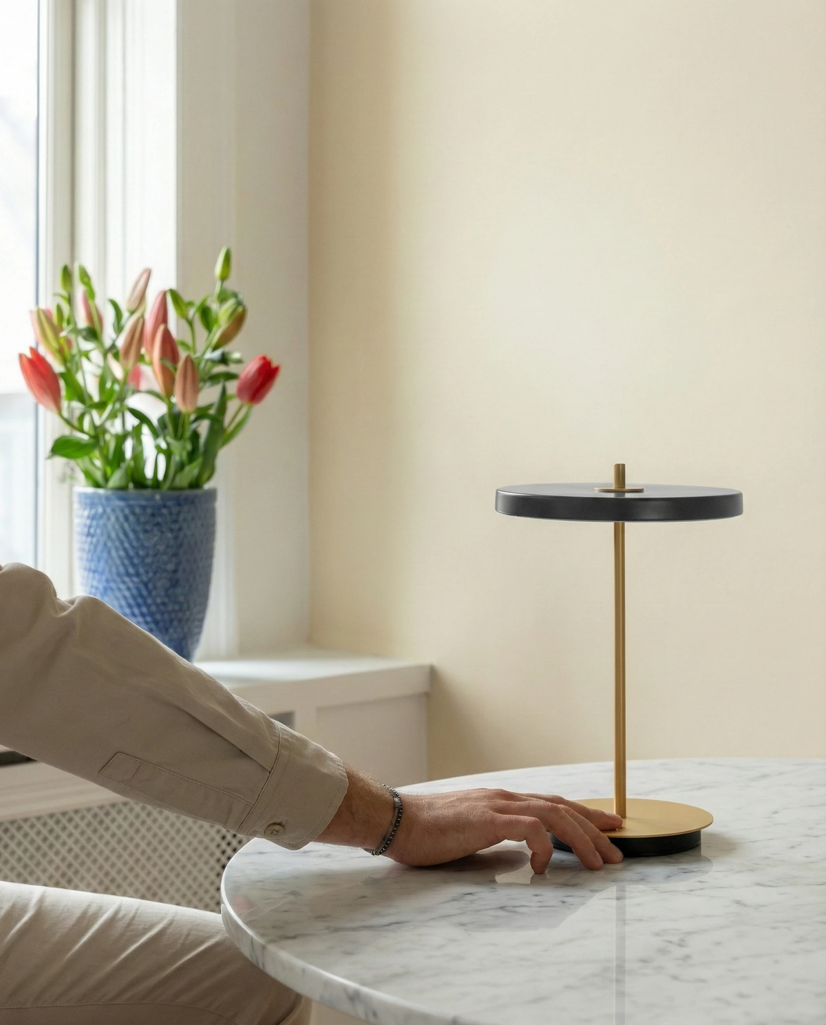 Cordless table lamp SALA No.01 in matte black on a marble table by a window with tulips, a hand touching the base