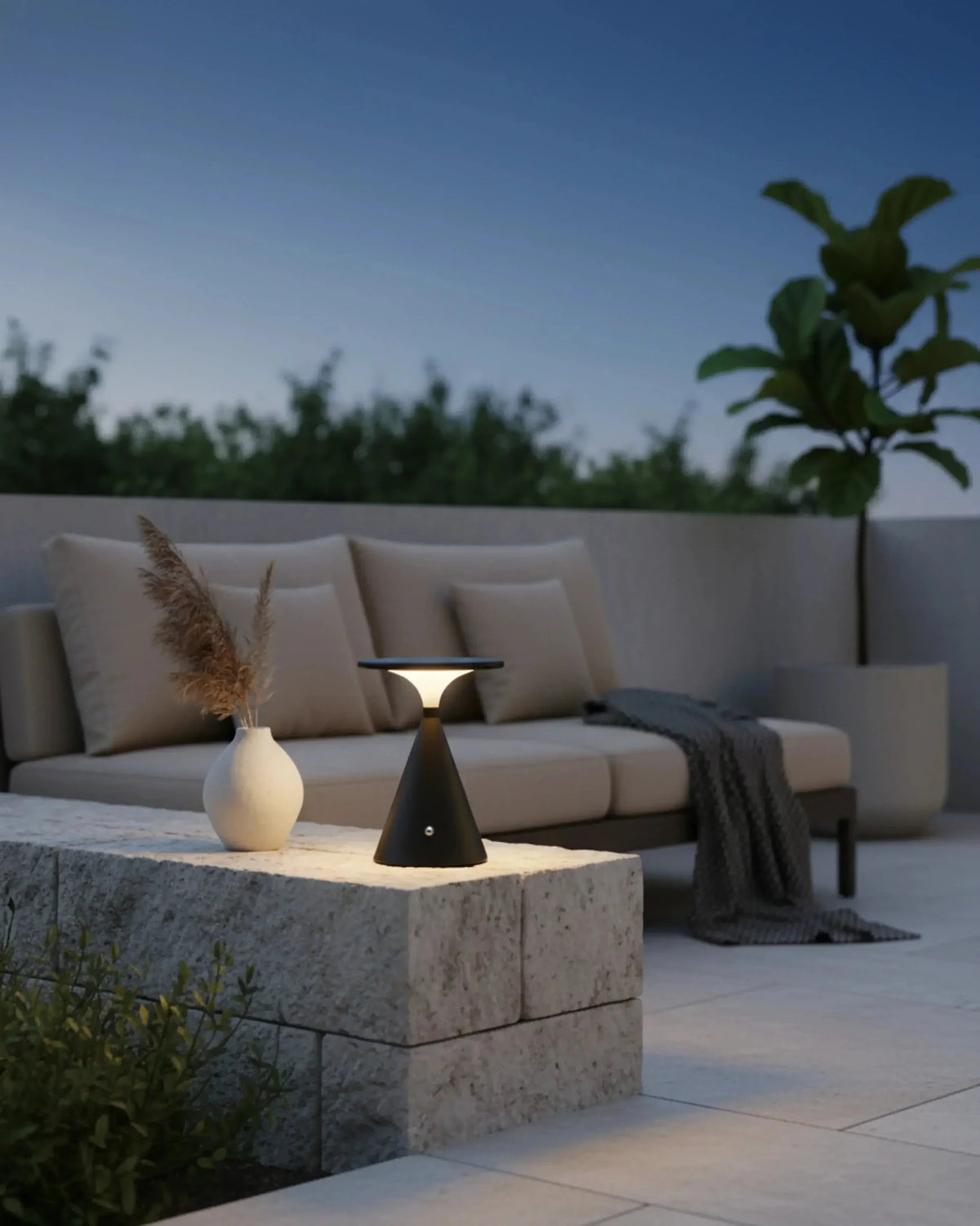Cordless table lamp SOSTA No.01 in black lit on a stone table on a patio at dusk with an outdoor sofa