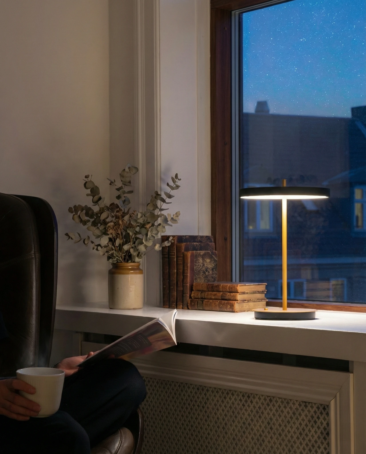 Cordless table lamp SALA No.01 in matte black, lit at night on a windowsill beside a reader with a mug