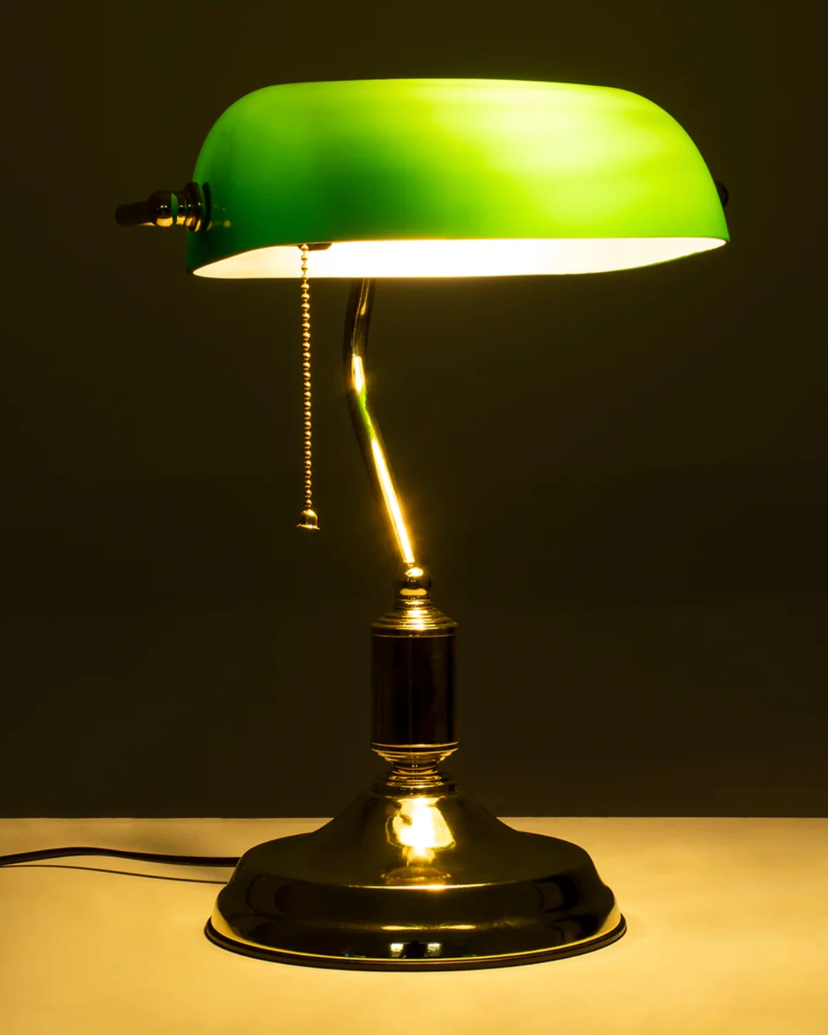 Glass desk lamp BANCO No.01, green glass shade glowing brightly over a dark base with pull chain, switched on
