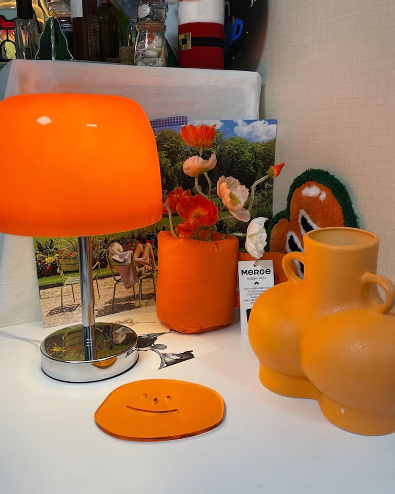 Glass table lamp VENEZIA No.01 switched on, orange shade glowing among orange vases and a smiley coaster