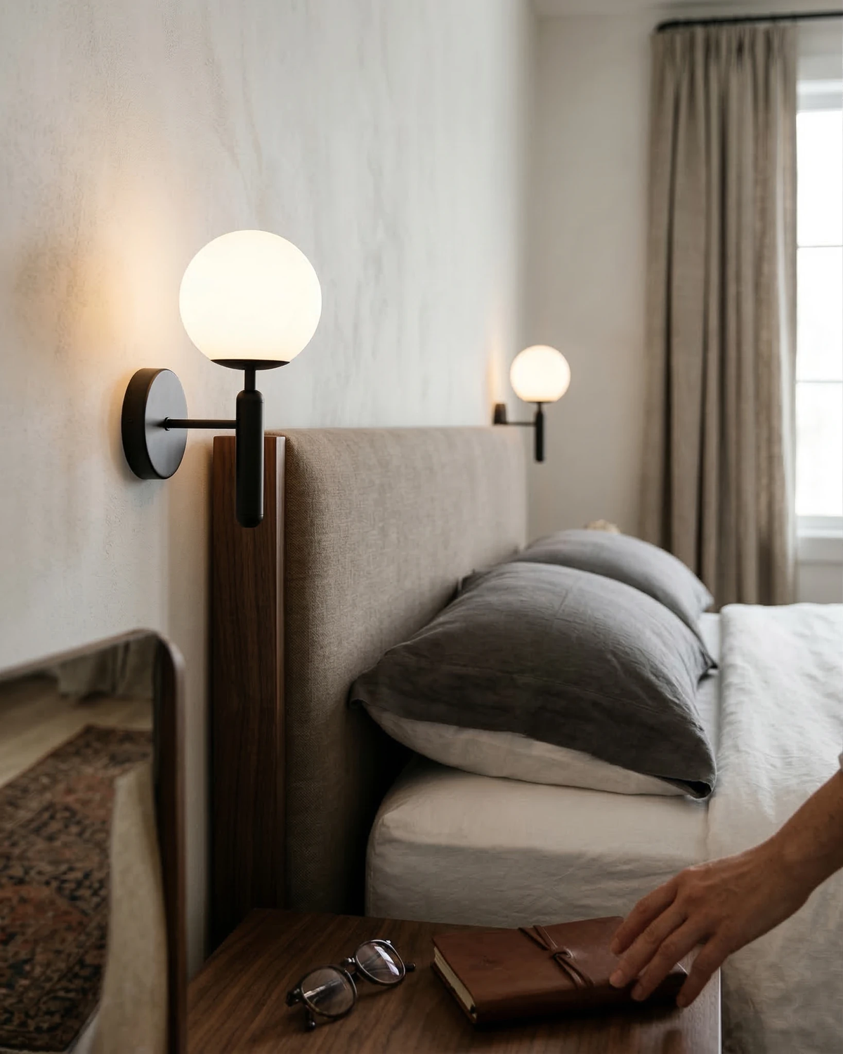 Two globe wall sconces CIELO No.01 in black lit beside a bed as a hand sets glasses on the nightstand