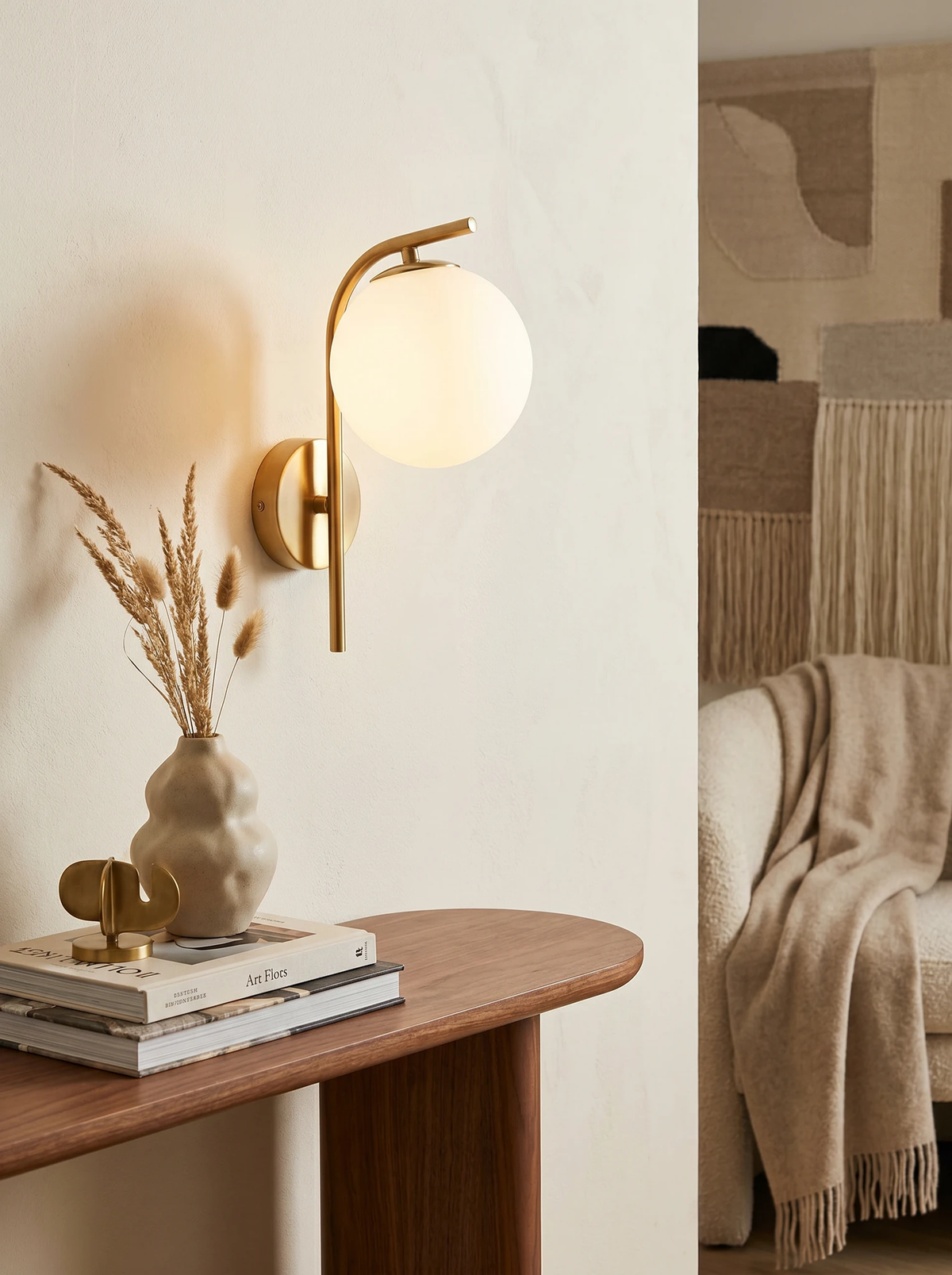 Globe wall sconce MIRA No.01 in brass lit on a plaster wall above a walnut console with books and a vase