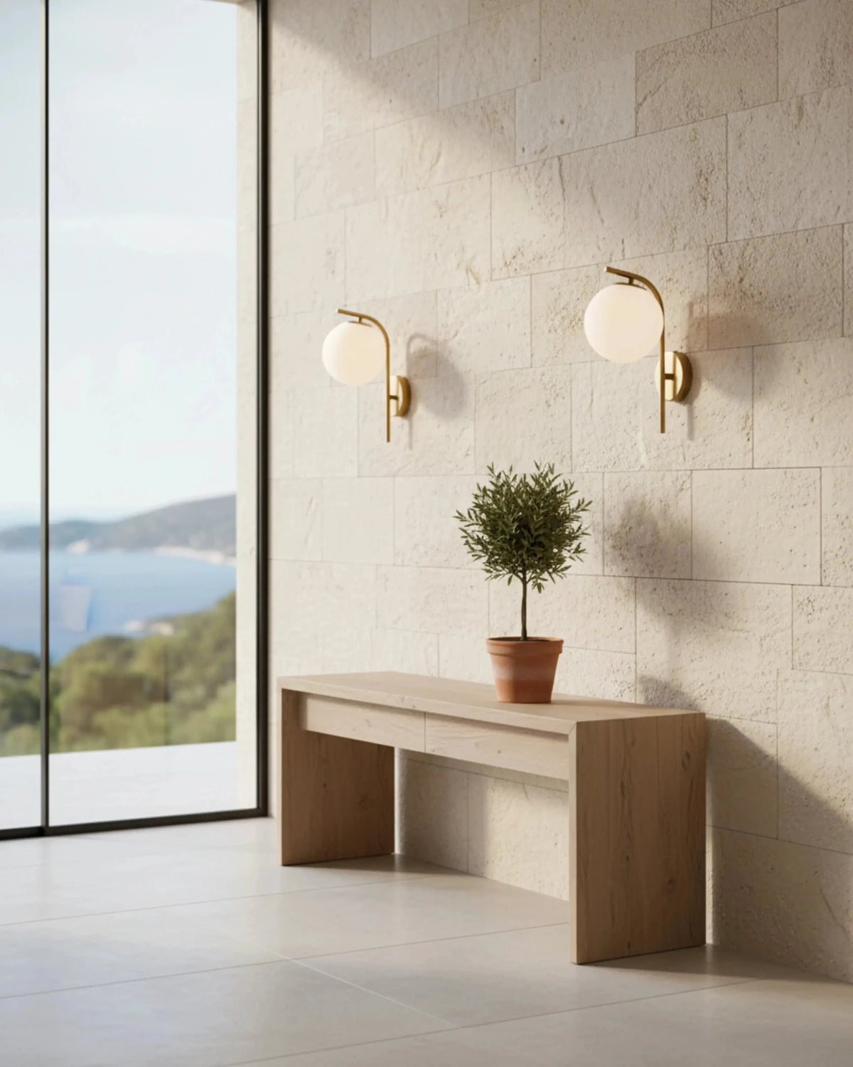 Two globe wall sconces MIRA No.01 in brass on a travertine tiled wall above a wood bench with an olive tree