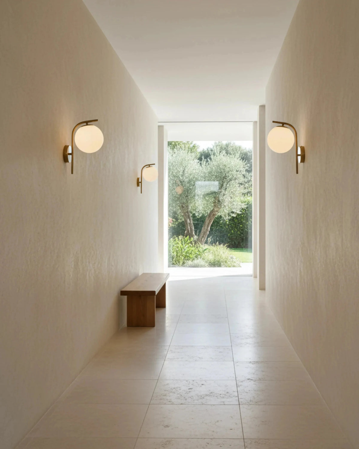 Globe wall sconces MIRA No.01 in brass along both walls of a plaster hallway opening onto a garden