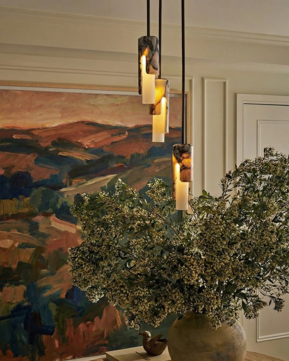 Three marble pendant lights PALERMO No.01 lit in a cluster beside a large landscape painting and a vase of greenery
