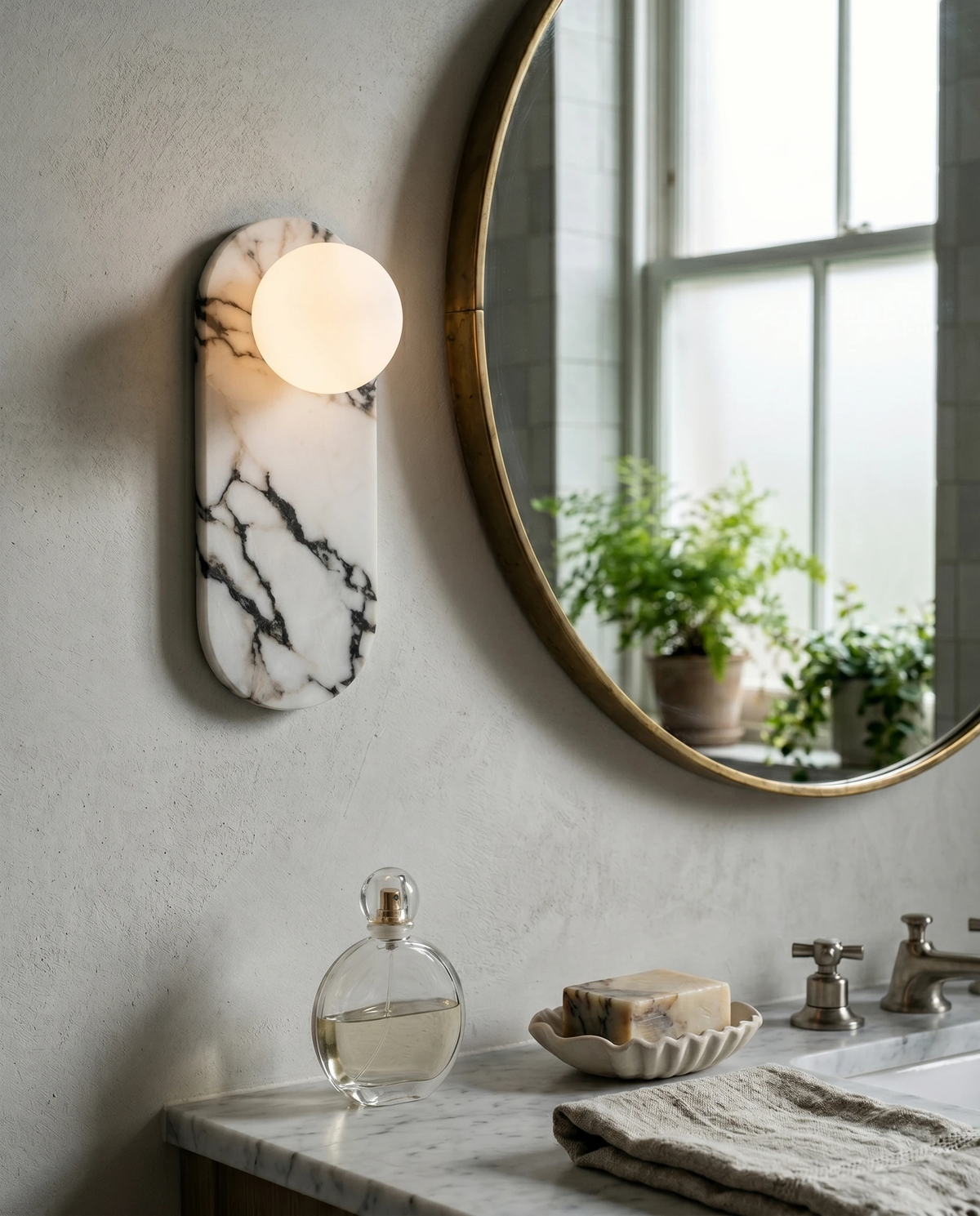 Marble wall sconce AVOLA No.01 lit beside a round brass mirror above a bathroom sink with plants