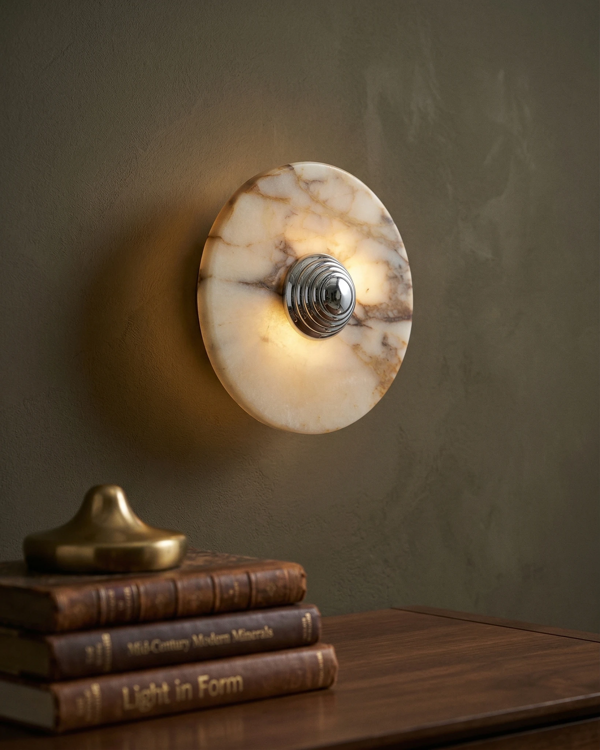 Marble wall sconce LATTE No.01 with chrome center lit on a dark plaster wall above a wooden desk with stacked books