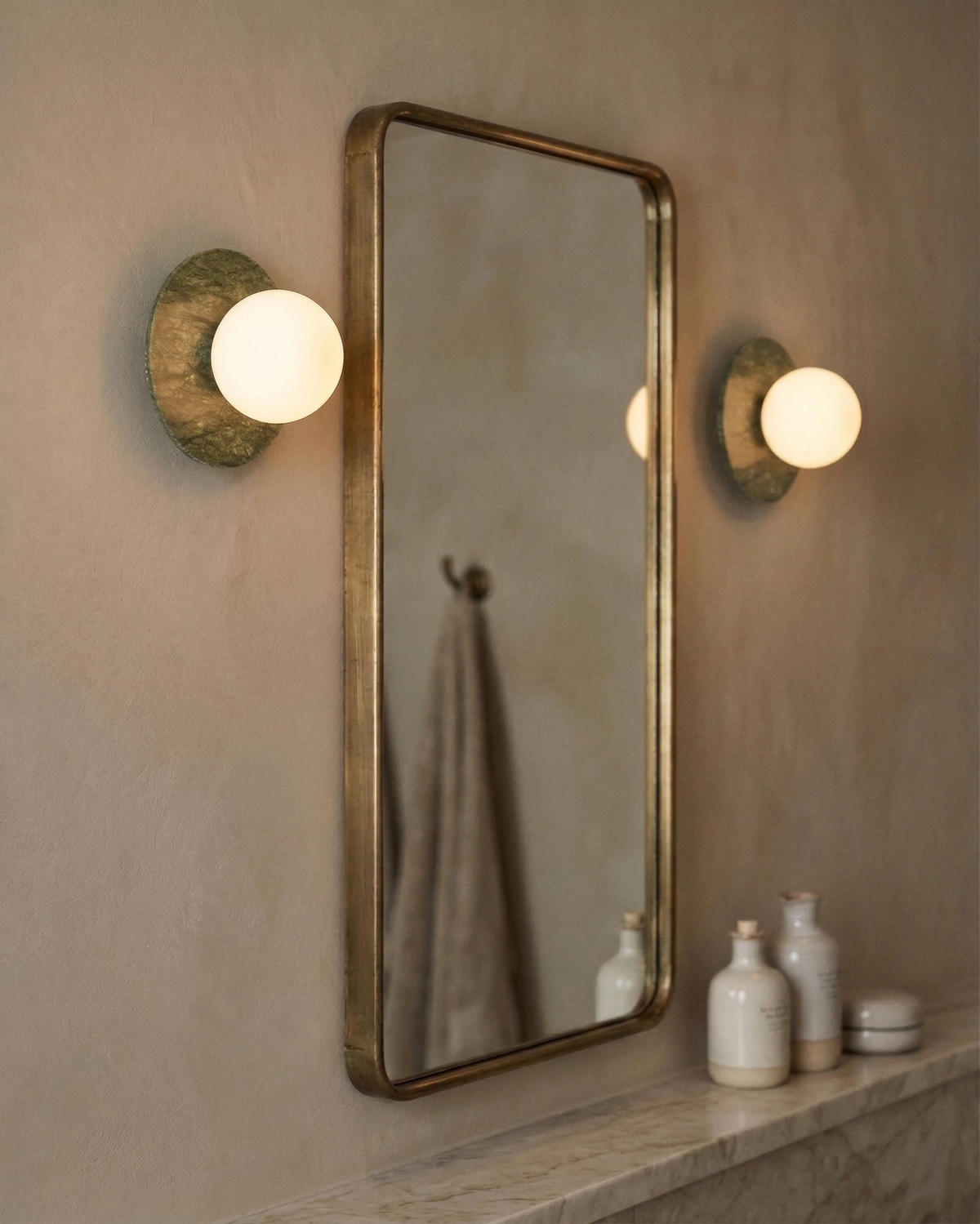 Pair of marble wall sconces VENA No.01 glowing beside a brass-framed mirror above a marble ledge with bottles