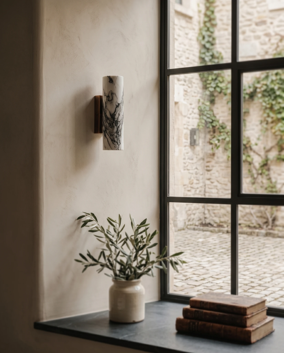 Marble wall sconce PORTORO No.01 on a plaster wall beside a steel-framed window with an olive plant and books