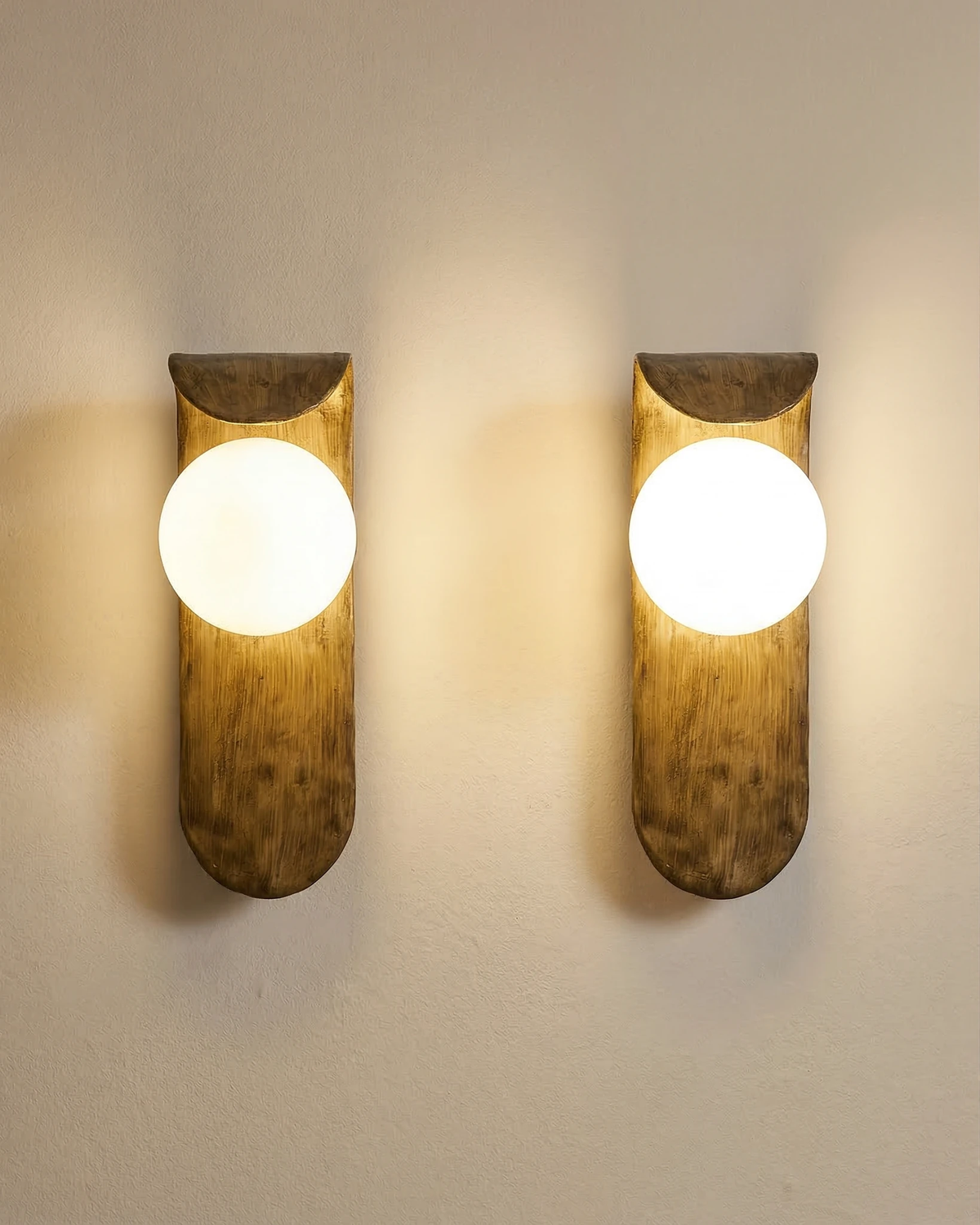 Pair of modern wall sconces ANIMA No.02 lit side by side on a beige wall, sculpted bodies and glowing globes