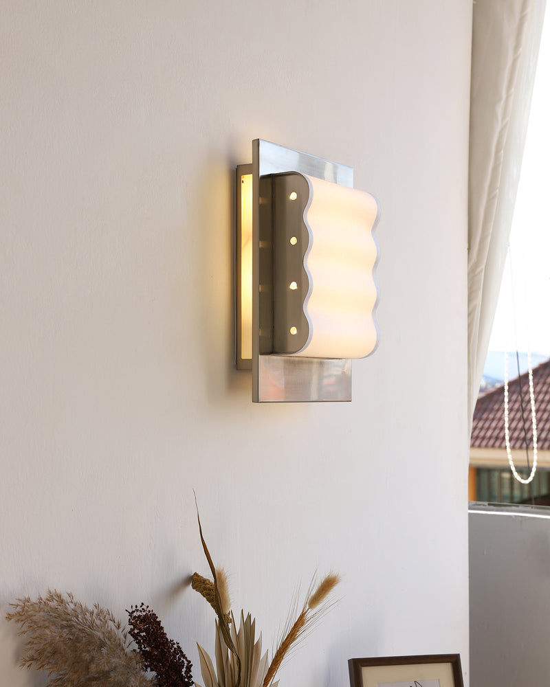 Modern wall sconce MILANO No.01 glowing on a white wall by a window above a vase of dried grasses and a frame