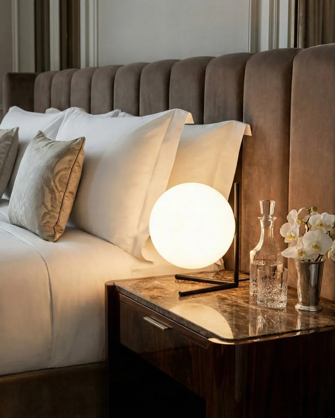 Opal globe table lamp ARIA No.01 glowing on a nightstand next to a bed with a channel-tufted headboard