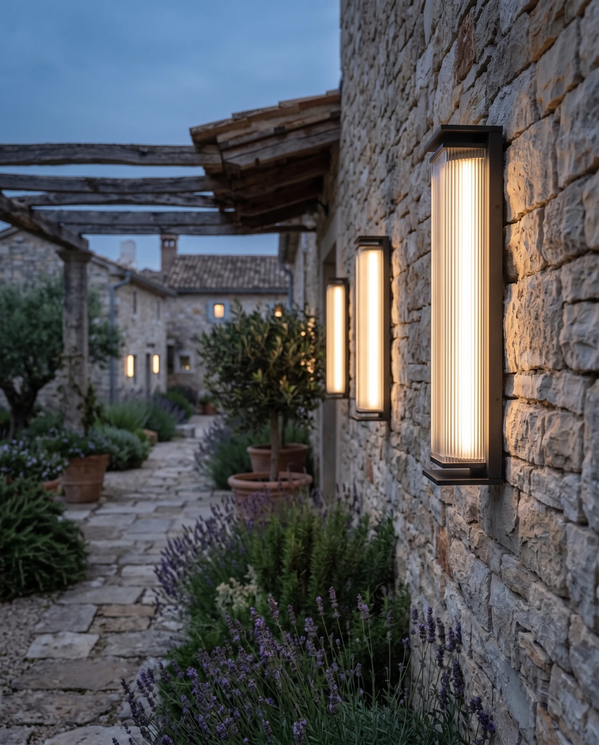 Outdoor wall sconce COSTA No.01, two units lit on a stone facade beside a garden path lined with lavender