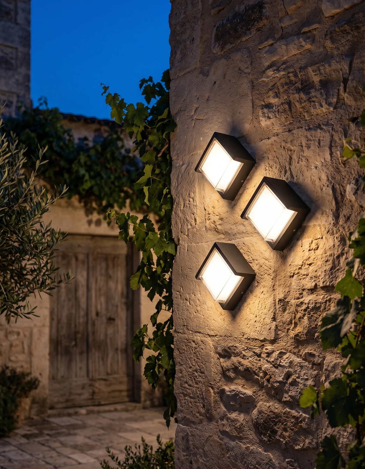Three outdoor wall sconces FARA No.02 lit on a stone wall with climbing vines at twilight