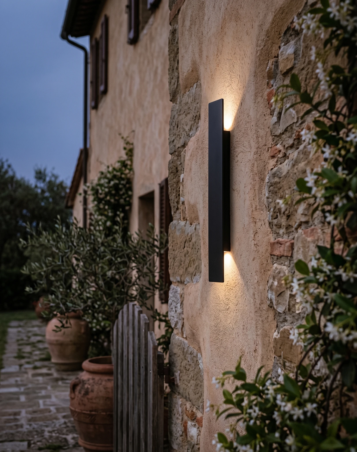 Outdoor wall sconce COLLE No.01 in matte black lit up and down on a stone farmhouse facade at dusk