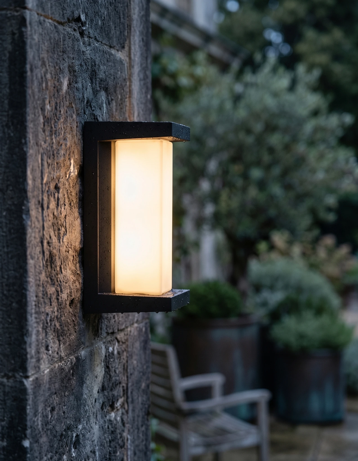 Outdoor wall sconce PORTICO No.02 switched on against a rough stone wall with a garden bench and planters