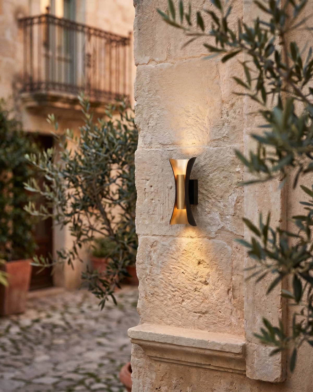 Outdoor wall sconce VENTO No.01 lit on a limestone wall in a cobbled courtyard with olive trees and a balcony