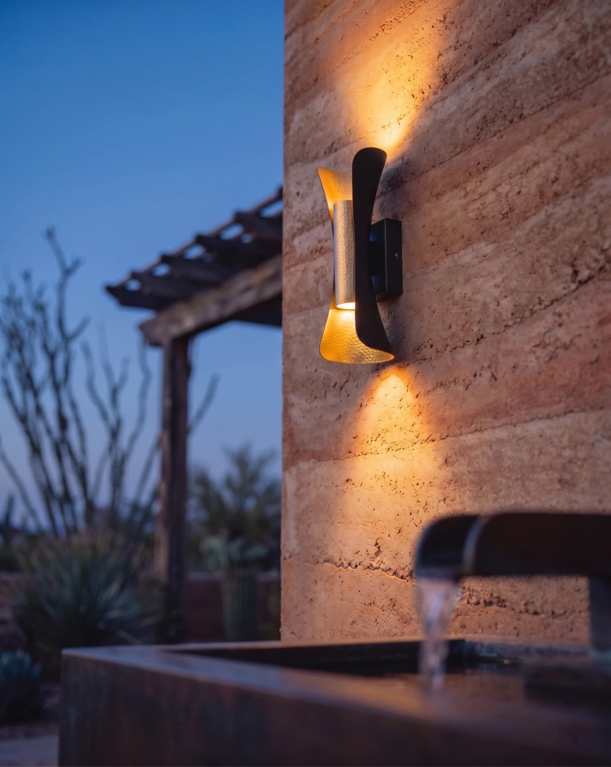 Outdoor wall sconce VENTO No.01 lit on a rammed-earth wall at twilight with desert plants and a pergola behind