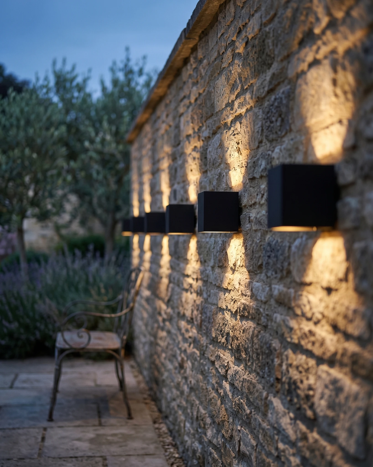Three outdoor wall sconces FARA No.01 in black lit along a stone garden wall with olive trees at dusk