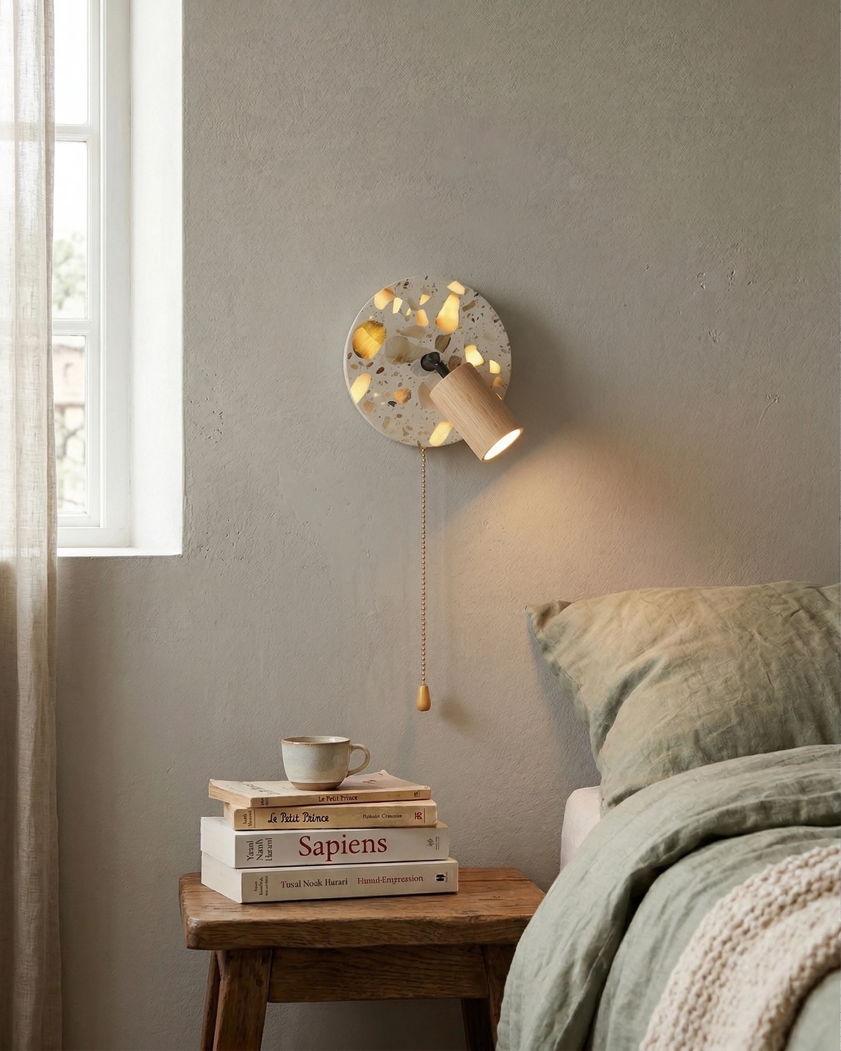 Terrazzo wall sconce PIETRA No.08 in natural ash switched on above a rustic nightstand stacked with books