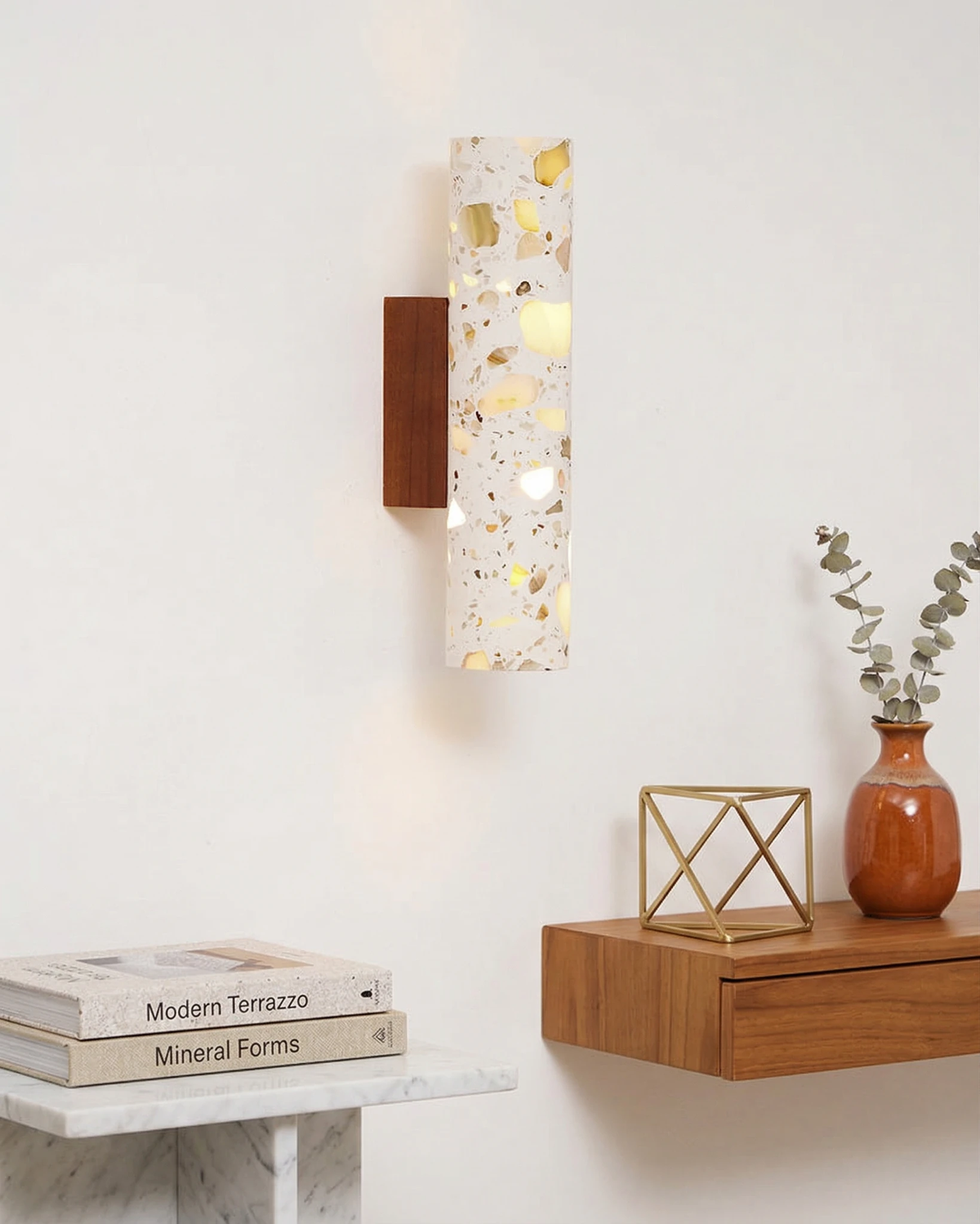 Terrazzo wall sconce INCA No.02 switched on above a floating wood shelf with books and a small vase