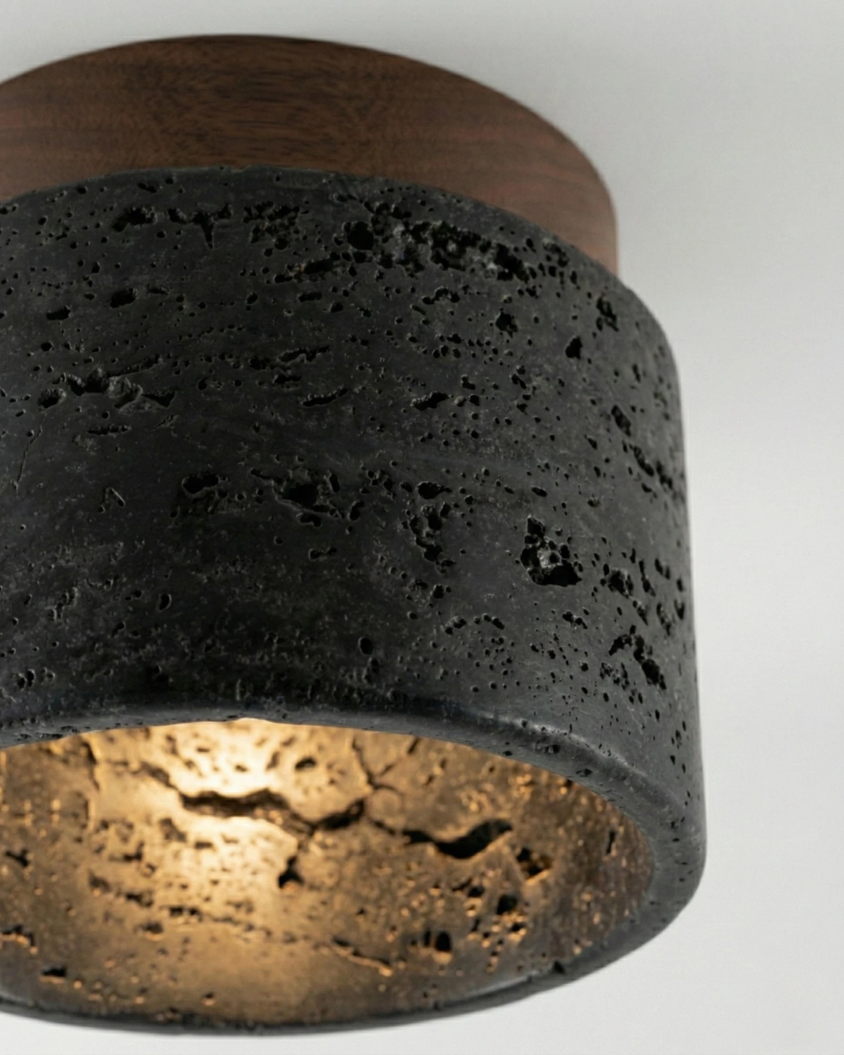 Close-up of the black travertine flush mount ceiling light VERTIN No.05 switched on, warm glow inside the pitted drum