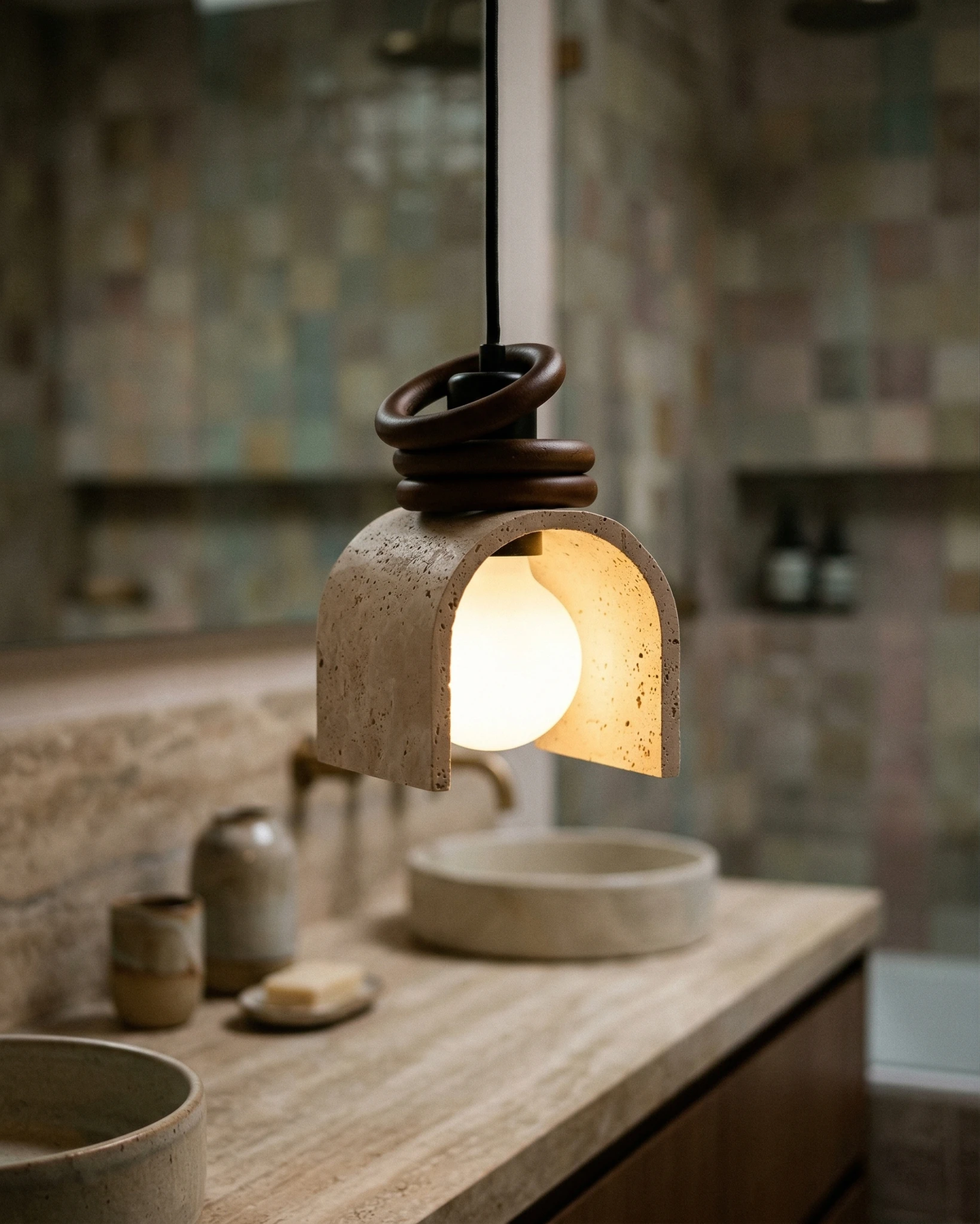Travertine pendant light ARCA No.02 lit above a travertine bathroom counter with a stone vessel sink and zellige tile