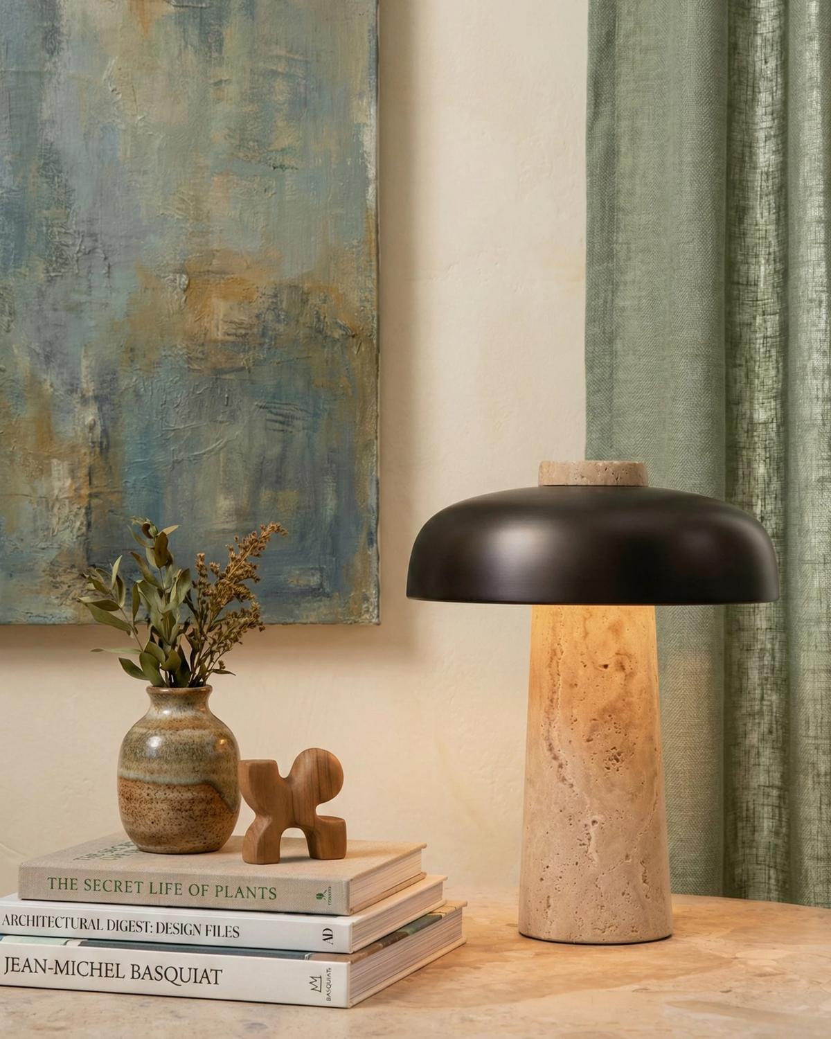 Travertine table lamp ROCCA No.03 lit on a console with stacked books and a vase beneath an abstract painting
