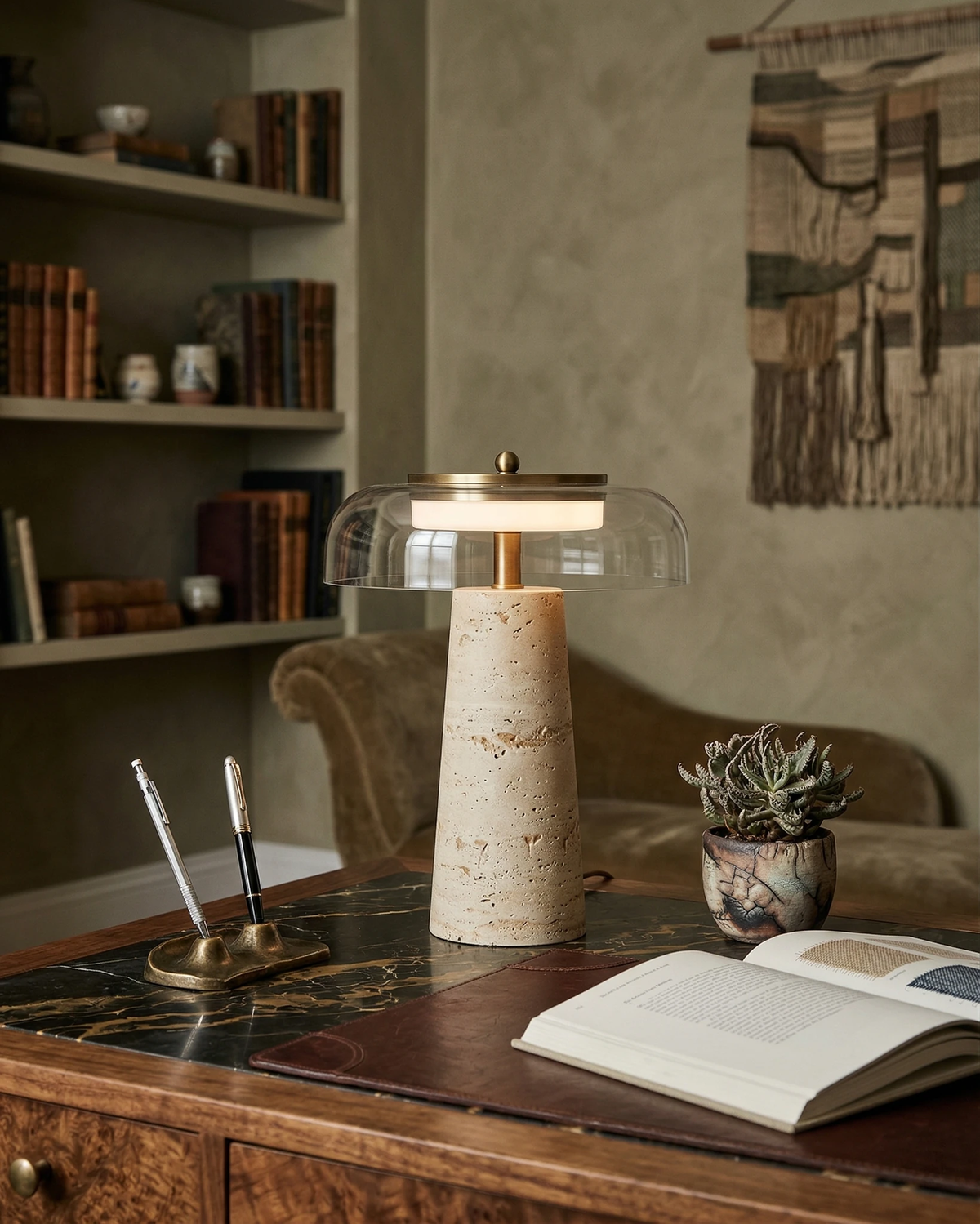Travertine table lamp MORA No.01 switched on atop a marble-topped wooden desk with an open book and shelves of books