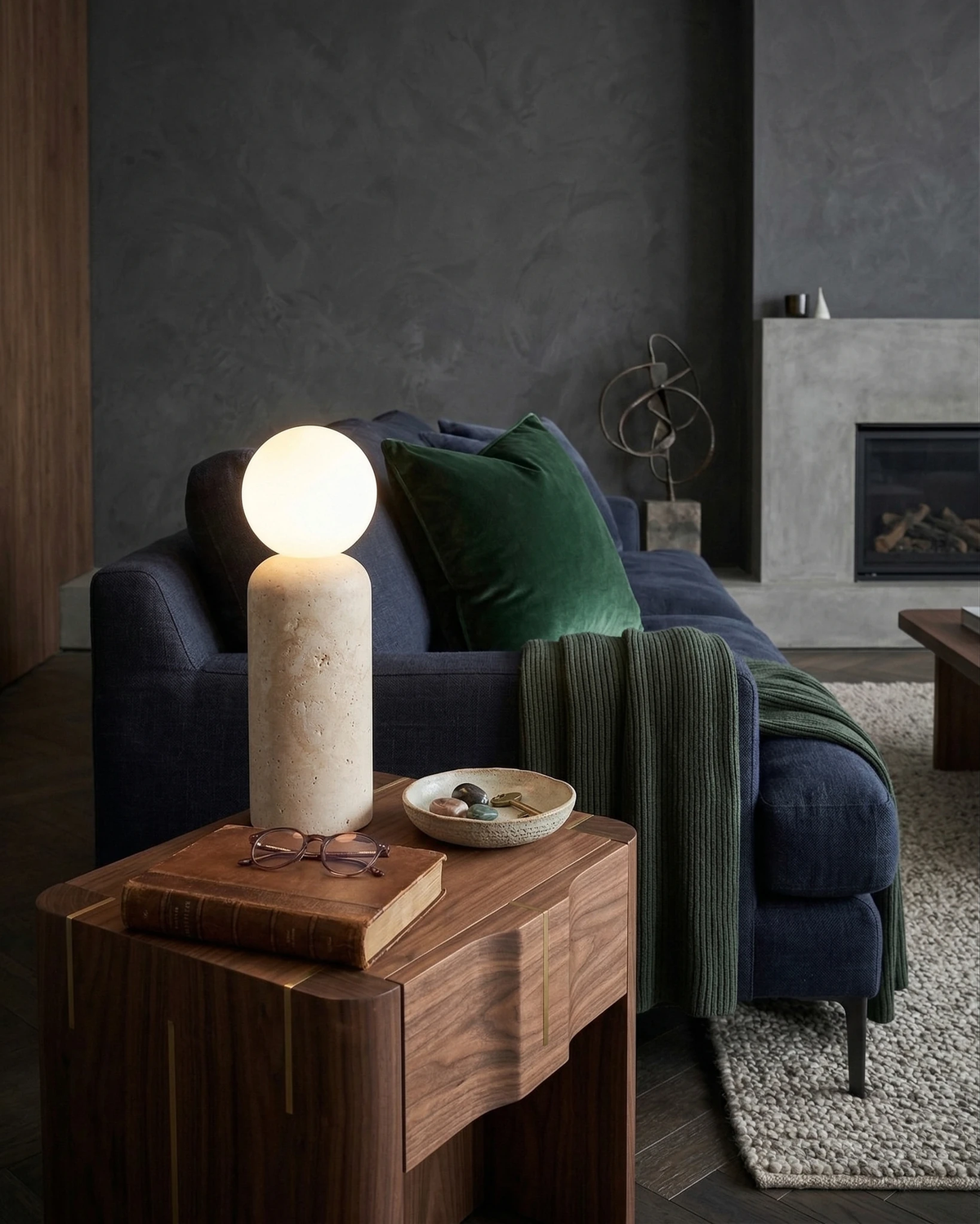 Travertine table lamp MORA No.04 cylindrical lit on a walnut side table next to a navy sofa and a concrete fireplace