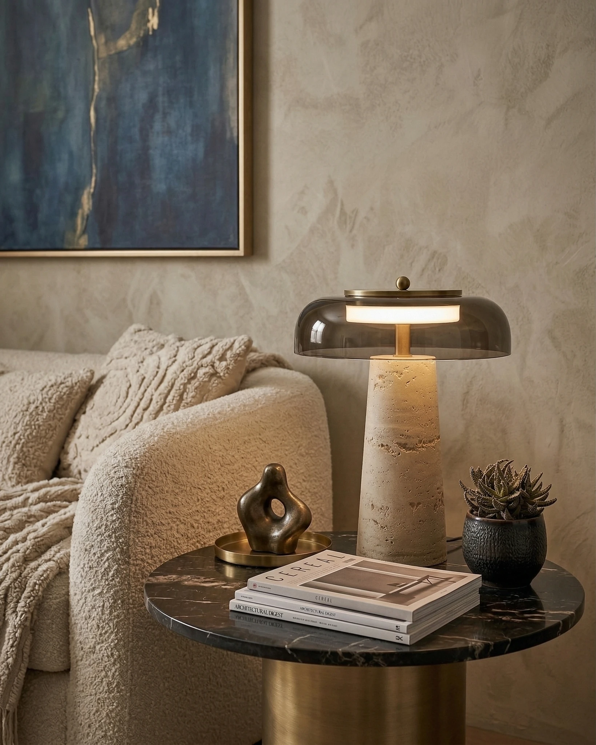 Travertine table lamp MORA No.01 with a smoky glass dome lit on a black marble side table next to a boucle sofa