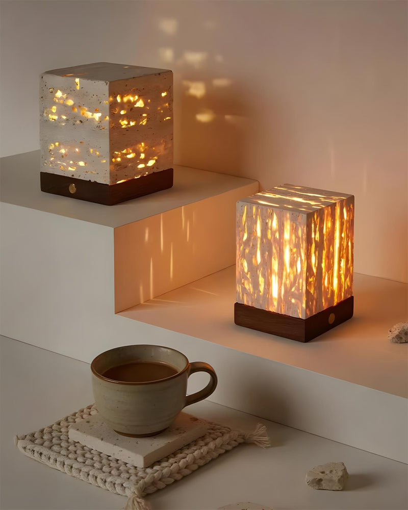Two travertine table lamps MORA No.05, narrow and wide cubes lit on white stepped shelves next to a mug of coffee