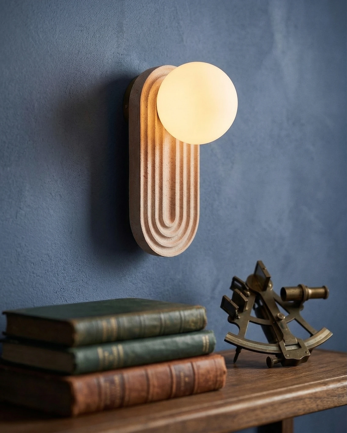 Travertine wall sconce ARCA No.01 lit on a deep blue plaster wall above antique books and a brass sextant