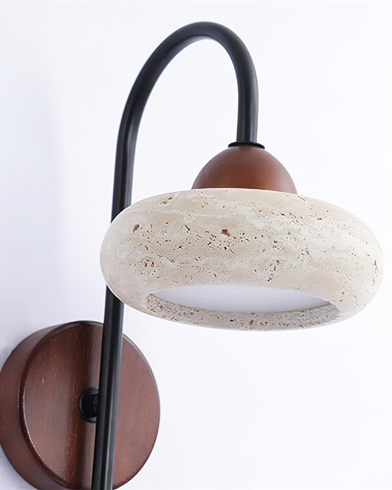 Travertine wall sconce BASSO No.01 seen up close, pitted stone donut under a walnut cap on the curved black arm, unlit