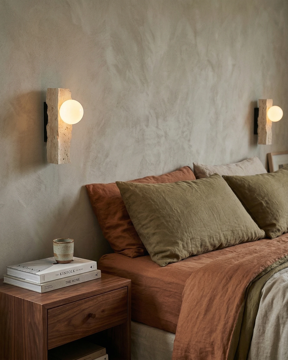 Two travertine wall sconces KANSO No.03 with lit opal globes flanking a bed dressed in rust and olive linen