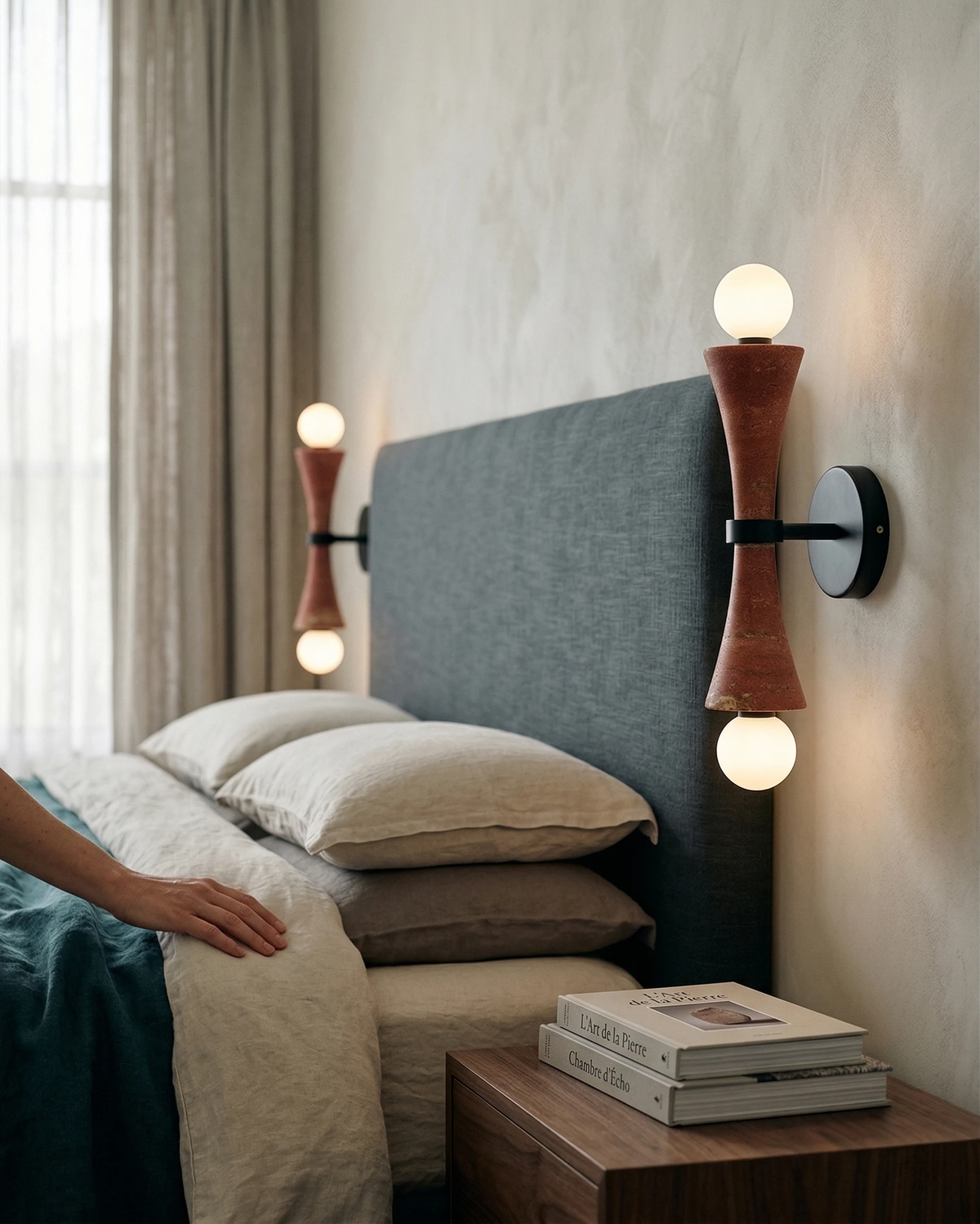 Travertine wall sconce VETTA No.01 in red travertine switched on beside a bed with a blue-gray headboard