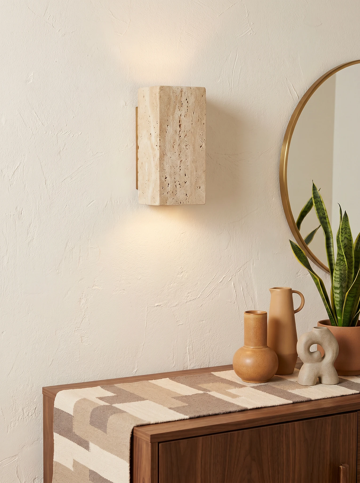 Travertine wall sconce GRANO No.01 lit above a walnut sideboard with ceramic vases and a round brass mirror