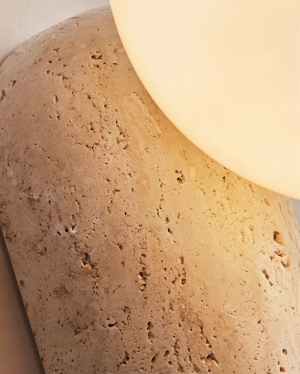 Close-up of the pitted travertine block and lit opal globe of the PERLA No.01 travertine wall sconce