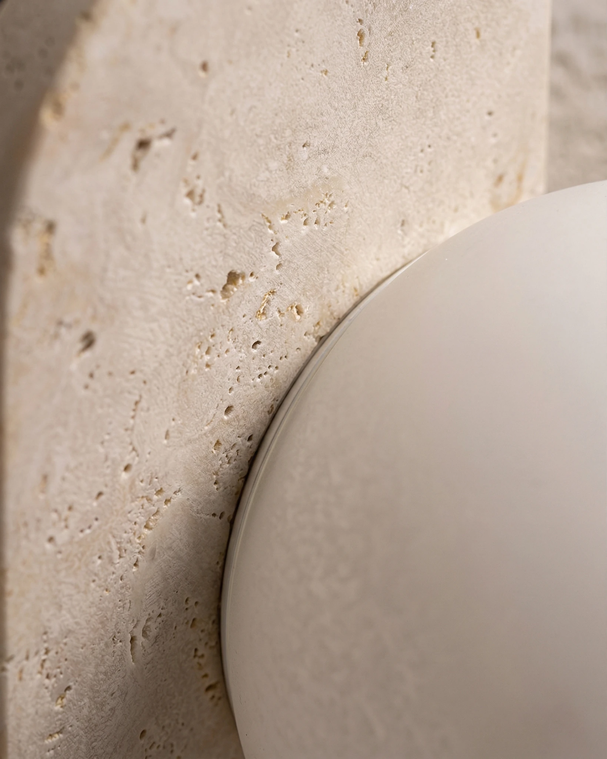 Close-up of the PRATO No.03 travertine wall sconce, opal globe against the pitted travertine plate