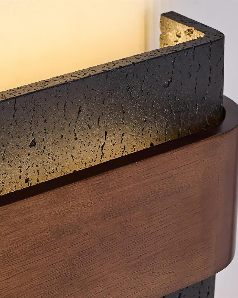 Close-up of the MASSO No.01 travertine wall sconce in Volcano Black, pitted dark stone above a curved walnut belt, lit