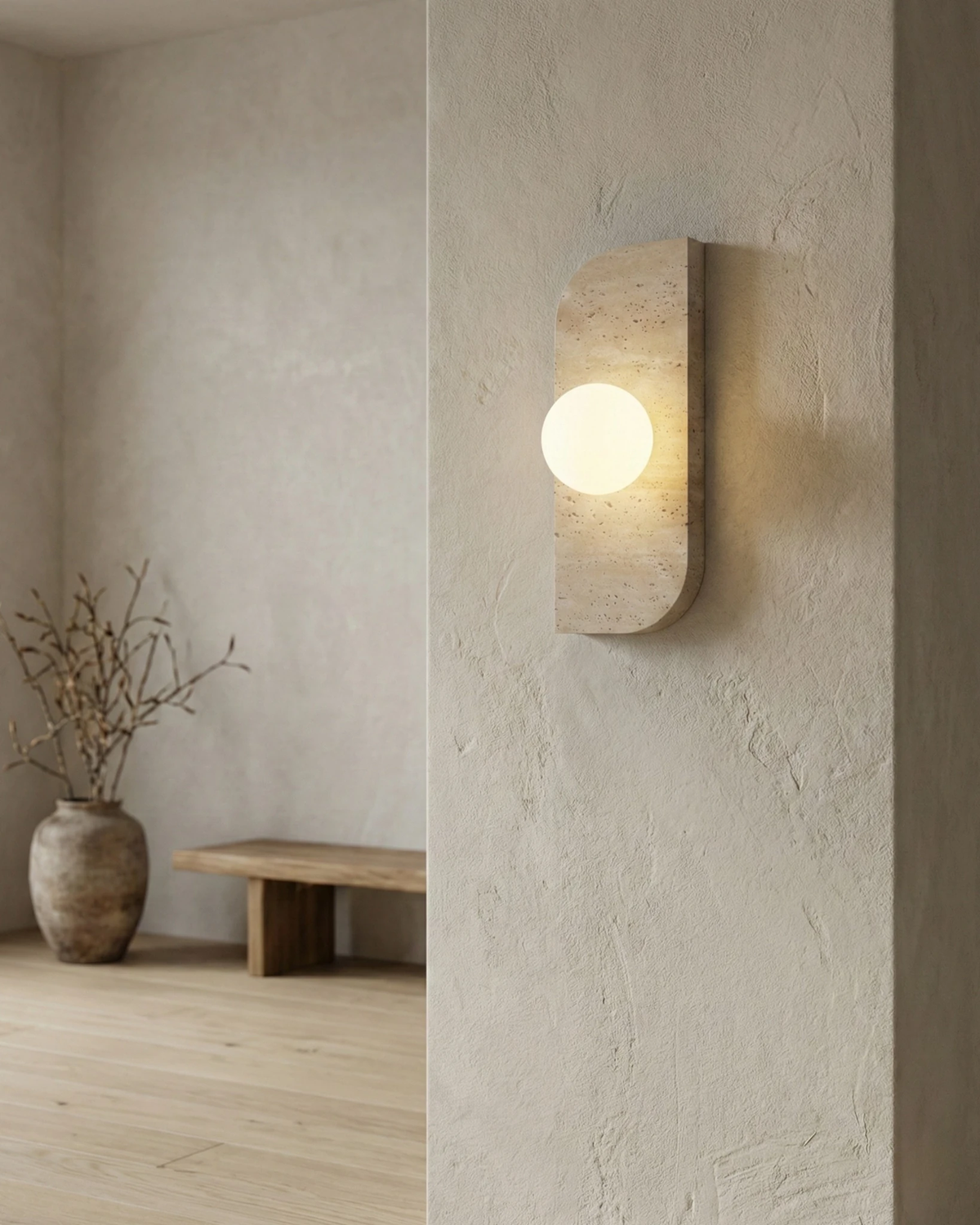 Travertine wall sconce GENOVA No.01 lit on a plaster partition in a minimal room with a wood bench and vase