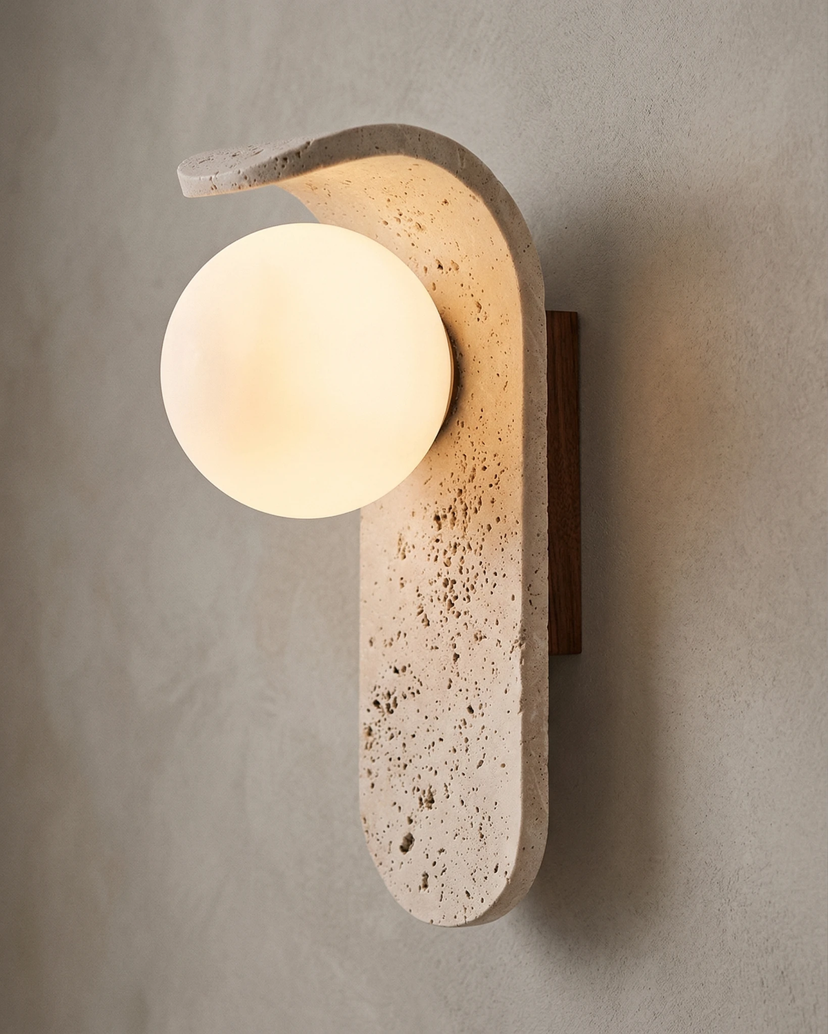 Travertine wall sconce ARCO No.01 lit on a plaster wall, opal globe glowing beneath the curved travertine