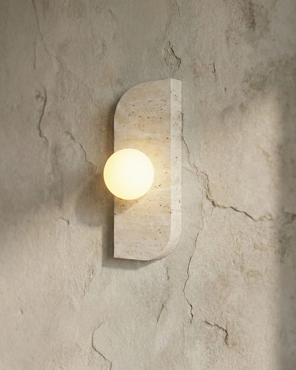 Travertine wall sconce GENOVA No.01 switched on against a cracked plaster wall, globe casting a warm glow